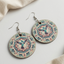 Round wooden drop earrings featuring a teal and pink hummingbird mandala design, resting flat on a light neutral background next to a soft linen cloth.