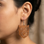 A detailed close-up photograph of a woman's ear wearing a large, intricately cut, warm brown wooden dangle earring with a stylized owl face and complex geometric patterns, hanging from a silver hook wire.