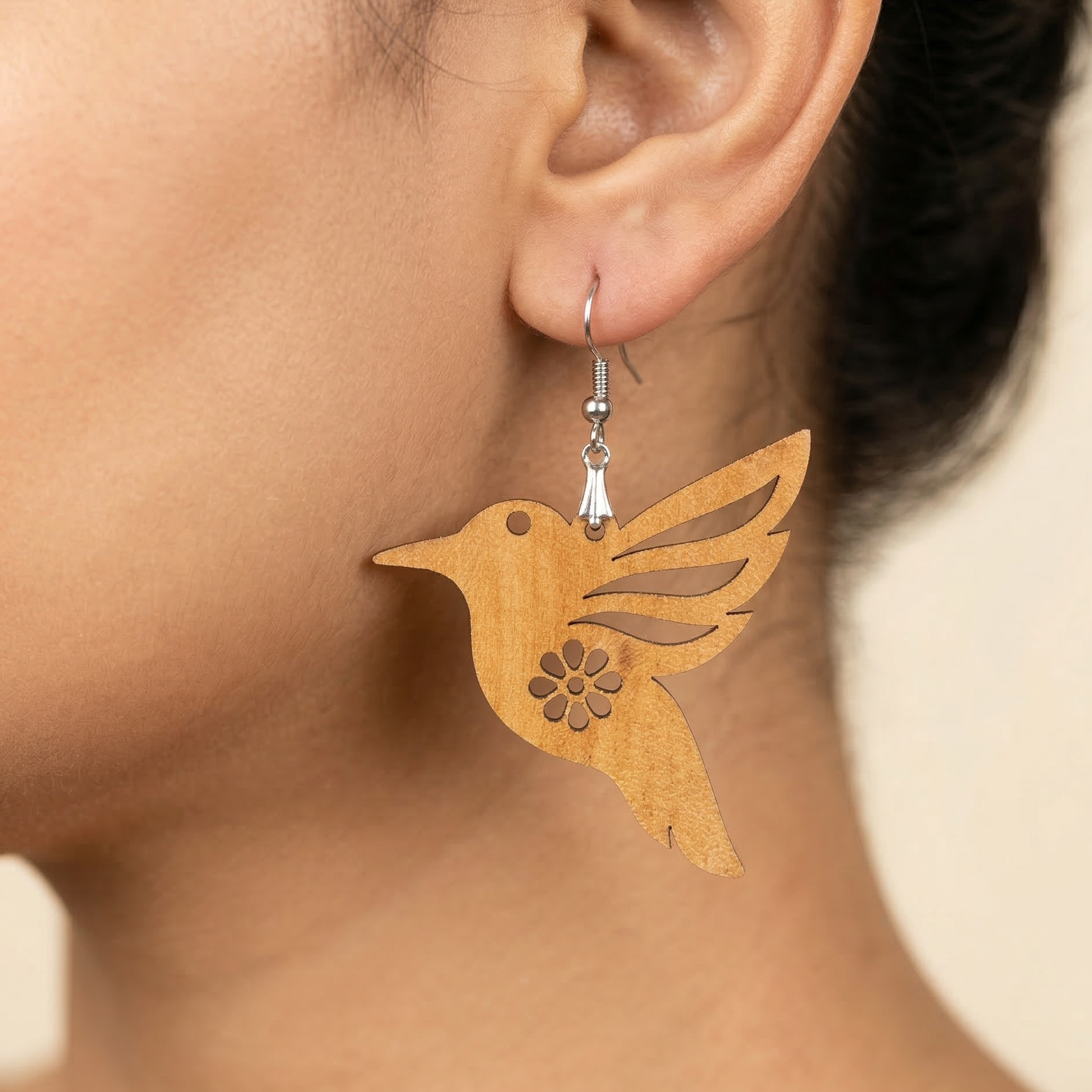 A detailed close-up photograph of a woman's ear wearing a delicate, laser-cut wooden hummingbird dangle earring with stylized, layered wings and a central eight-petal floral cutout, hanging from a silver hook, earth-toned and boho style. Main image