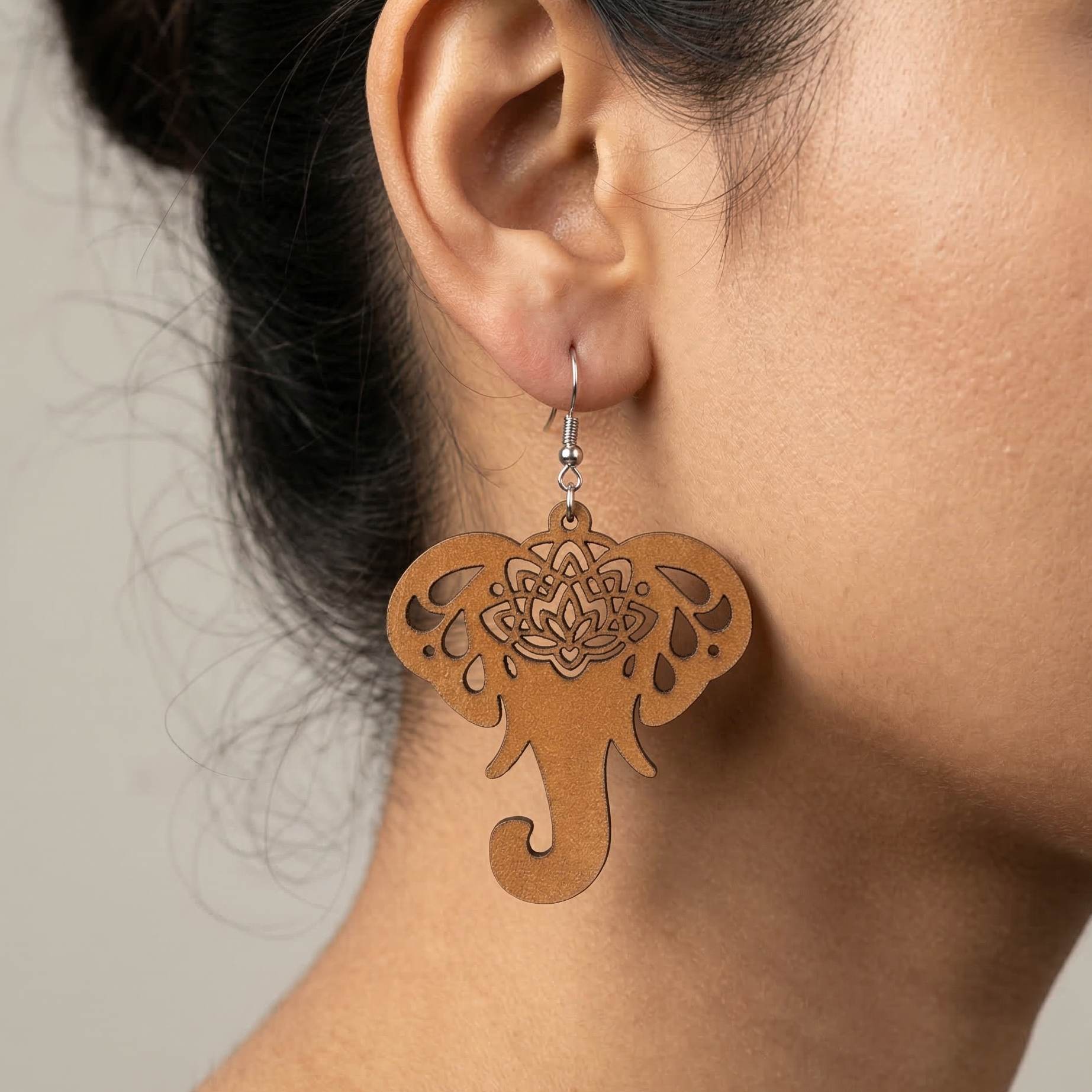 Light brown wooden elephant dangle earrings featuring an intricate laser-cut mandala design on the forehead, hanging from a silver metal hook, worn by a woman. Main image