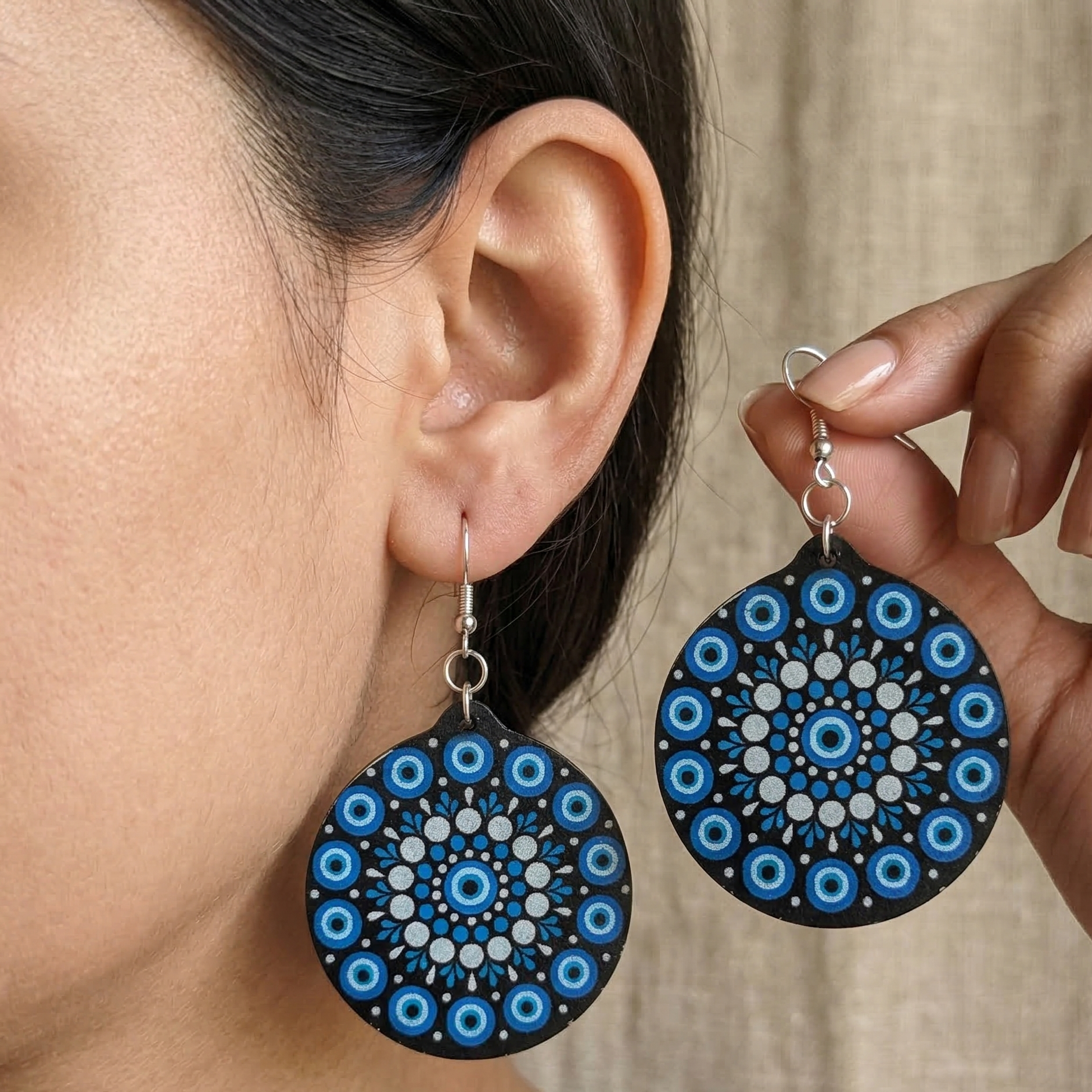 Woman wearing round wooden dangle earrings featuring an intricate blue, white, and black dot art mandala design. Main image