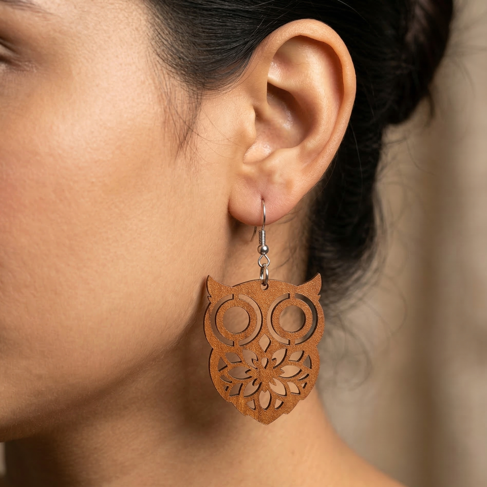 A detailed close-up photograph of a woman's ear wearing a large, intricately cut, warm brown wooden dangle earring with a stylized owl face and complex geometric patterns, hanging from a silver hook wire. Main image