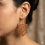 A detailed close-up photograph of a woman's ear wearing a large, intricately cut, warm brown wooden dangle earring with a stylized owl face and complex geometric patterns, hanging from a silver hook wire.