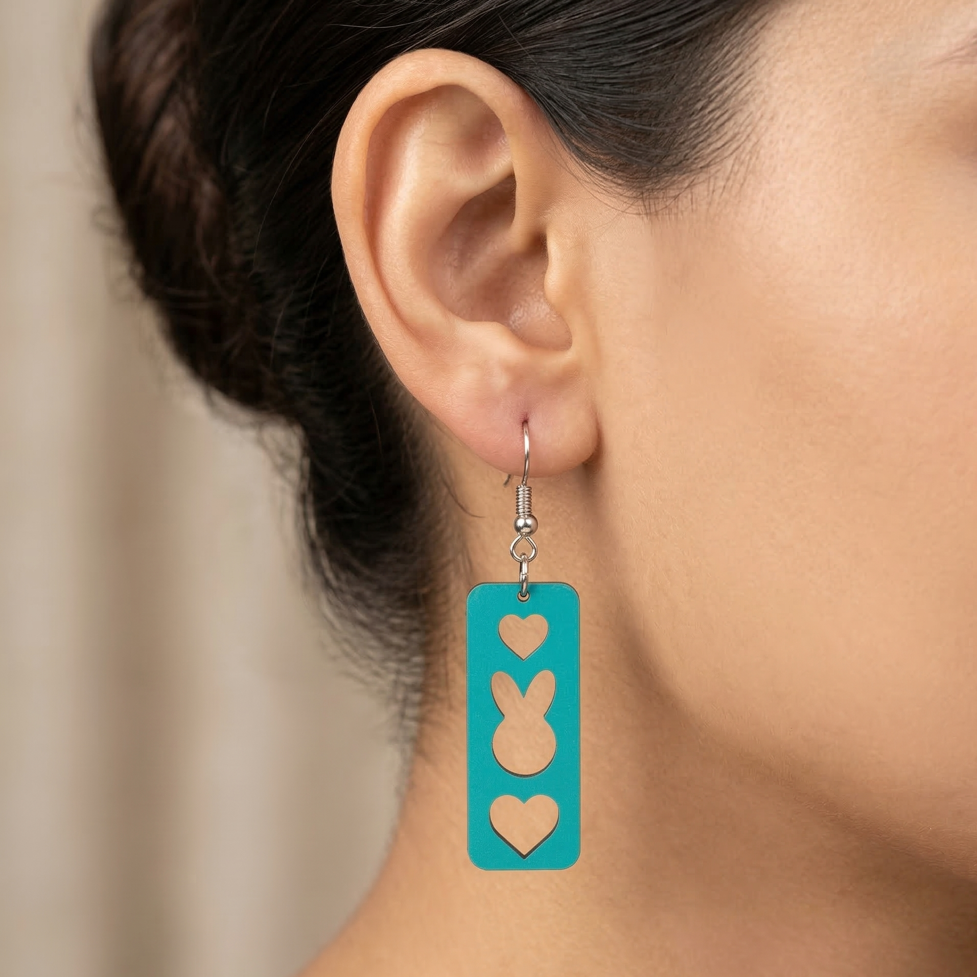 A detailed close-up photograph of a woman's ear wearing a teal-painted layered wooden dangle earring featuring distinct cutouts of a heart, a bunny face, and another heart, with natural wood grain visible in the cutouts, on a silver fishhook wire. Main image