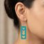 A detailed close-up photograph of a woman's ear wearing a teal-painted layered wooden dangle earring featuring distinct cutouts of a heart, a bunny face, and another heart, with natural wood grain visible in the cutouts, on a silver fishhook wire.