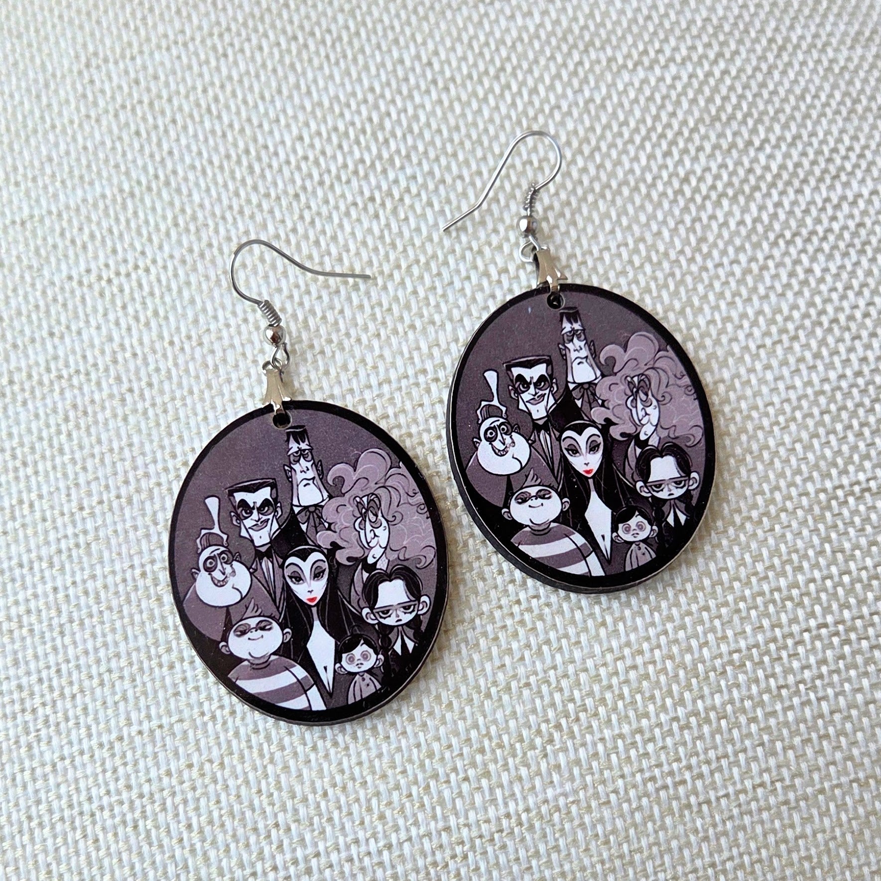 The Addams Family wooden earrings for women featuring Wednesday, Gomez, Morticia, Uncle Fester, Pugsley, Lurch, Grandmama, and the family in a monochrome oval design.